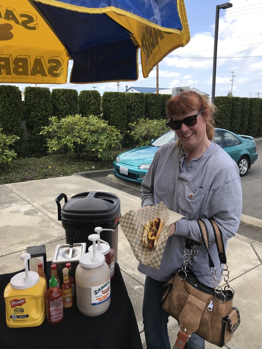 A woman holding her hotdog and smiling at the camera next to the "Housewife Hotdogs" cart